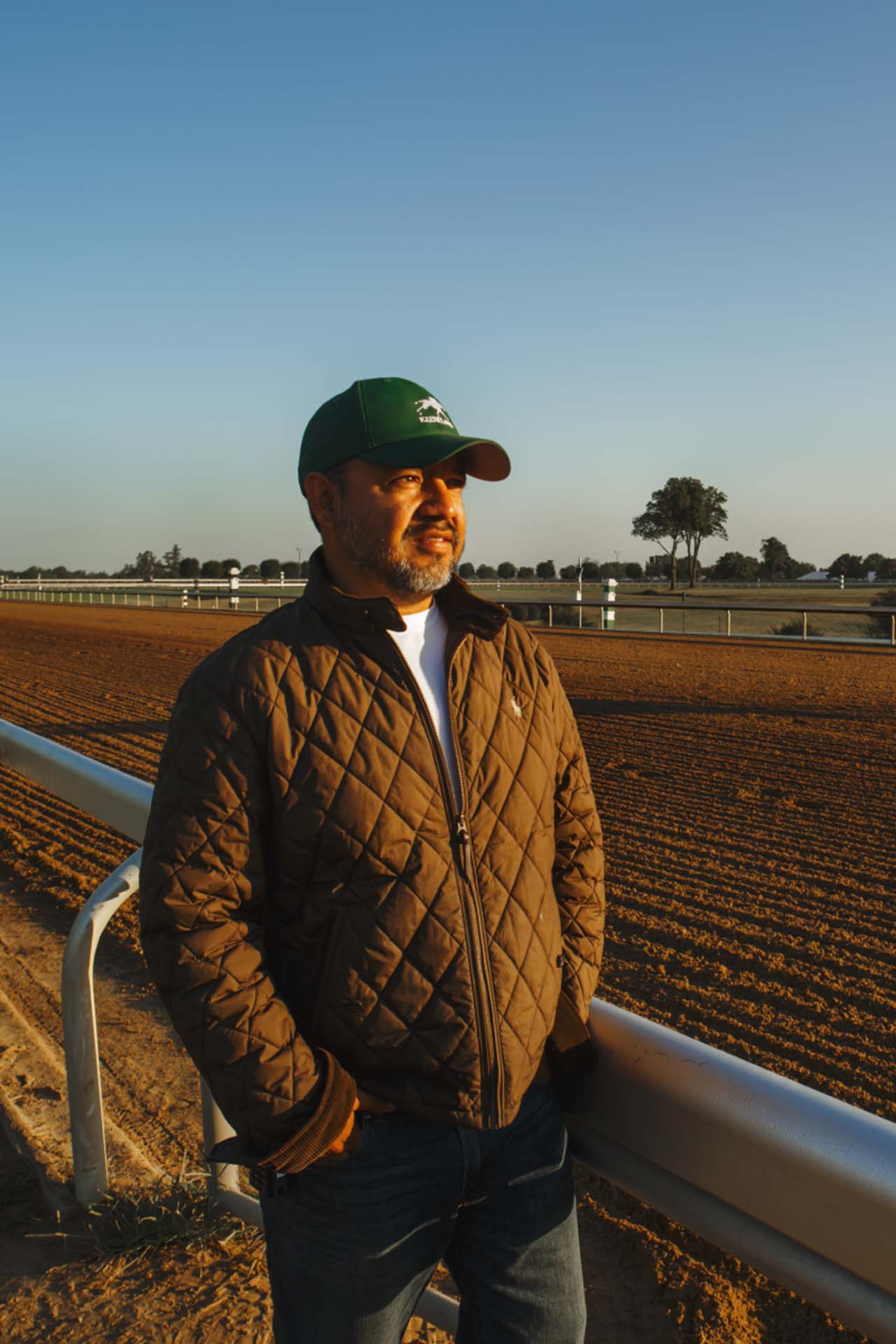 A photo of Alfredo Laureano looking off into the distance while standing by the dirt track at sunset. He is an older Hispanic man with graying chin stubble. He is wearing a green Keenlandsscape-brand hat and a brown Keenlandsscape-brand jacket overtop a white shirt.