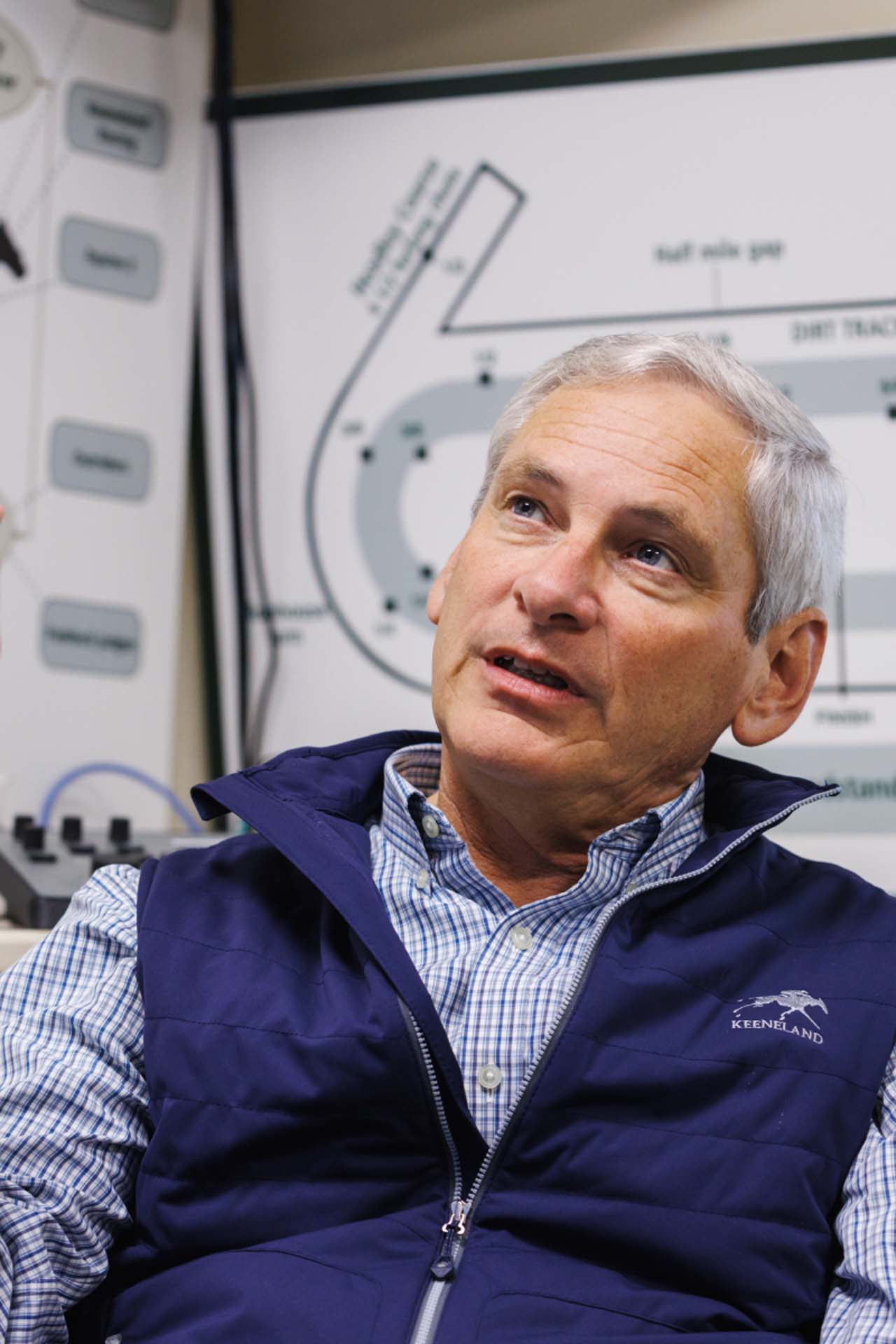 An up-close photo of Dr. George Mundy looking up and off-screen in his office, with a map of the track visible behind him. He is an older White man with short silver hair. He is wearing a navy Keenlandsscape-brand vest over a white and blue checkered shirt.